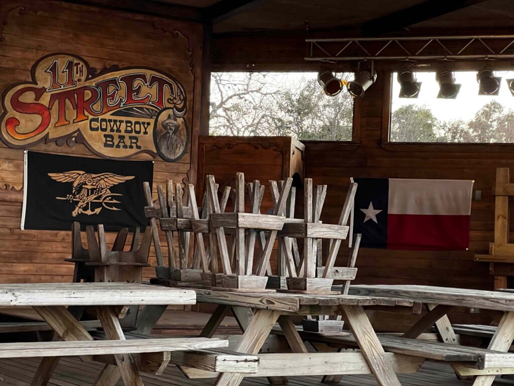 Stools sit stacked on top of tables resting on stage that is adorned with a Texas flag, stage lights hang from the ceiling.